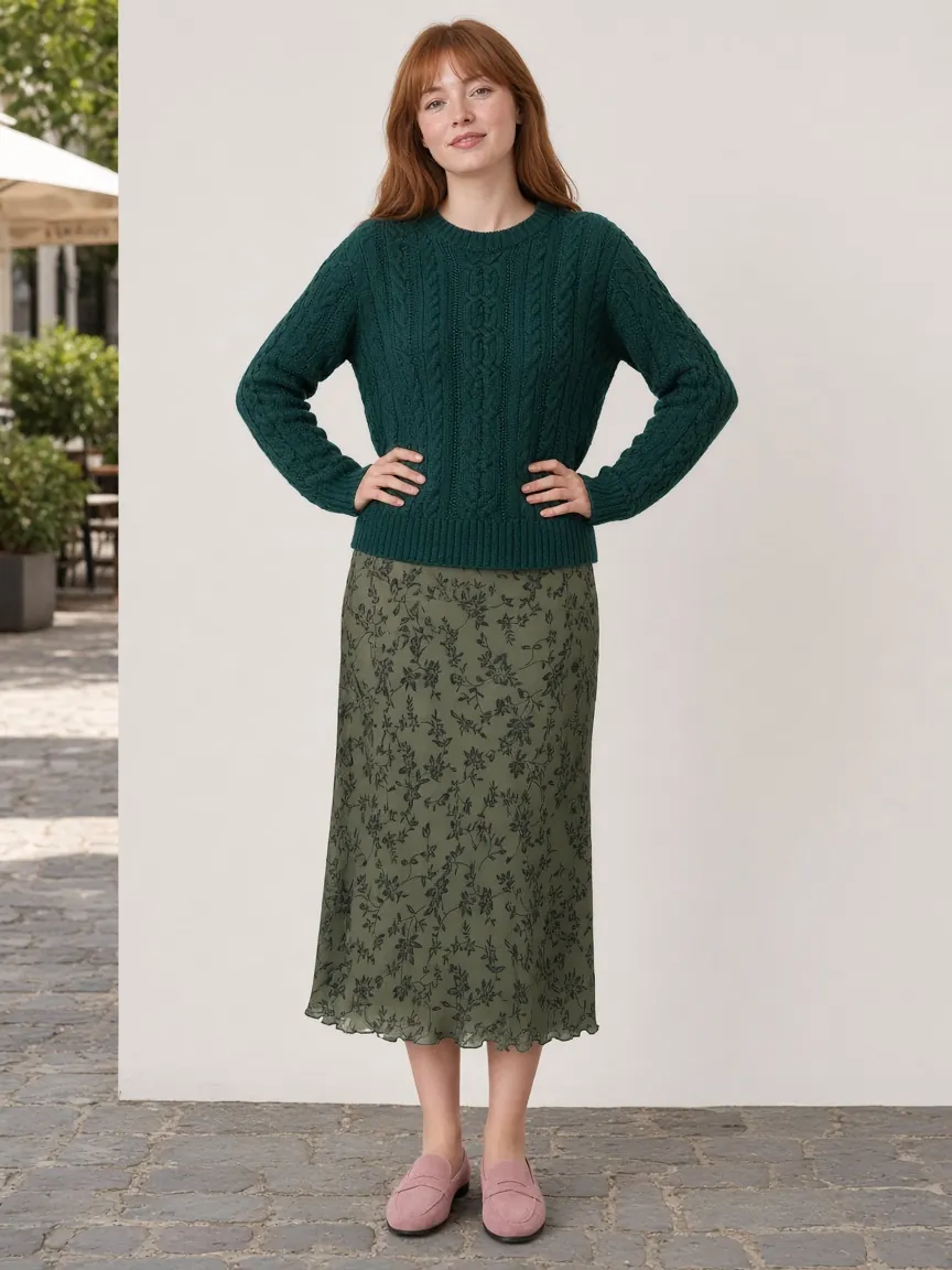 Woman in an emerald cable crewneck sweater worn untucked over a sage floral midi skirt and dusty-pink penny mules, standing front-on on a café sidewalk